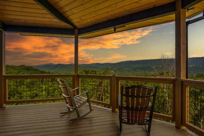 Cozy covered wooden deck with two rocking chairs overlooking green, forested mountains and a golden-orange sunset sky — panoramic mountain view.