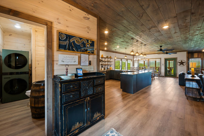 Open-concept rustic farmhouse interior with large kitchen island, pendant lights, wood-plank ceiling and floors, dining area, laundry nook and windows overlooking green countryside