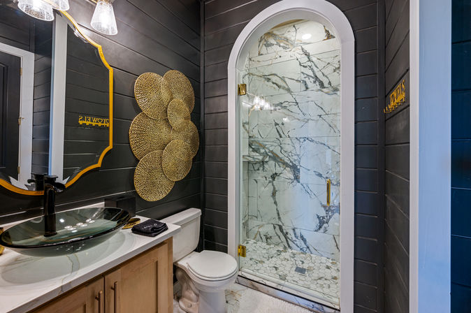 Modern bathroom with dark shiplap walls, arched glass marble-tiled shower, glass vessel sink on a white vanity, toilet, and round gold decorative accents.