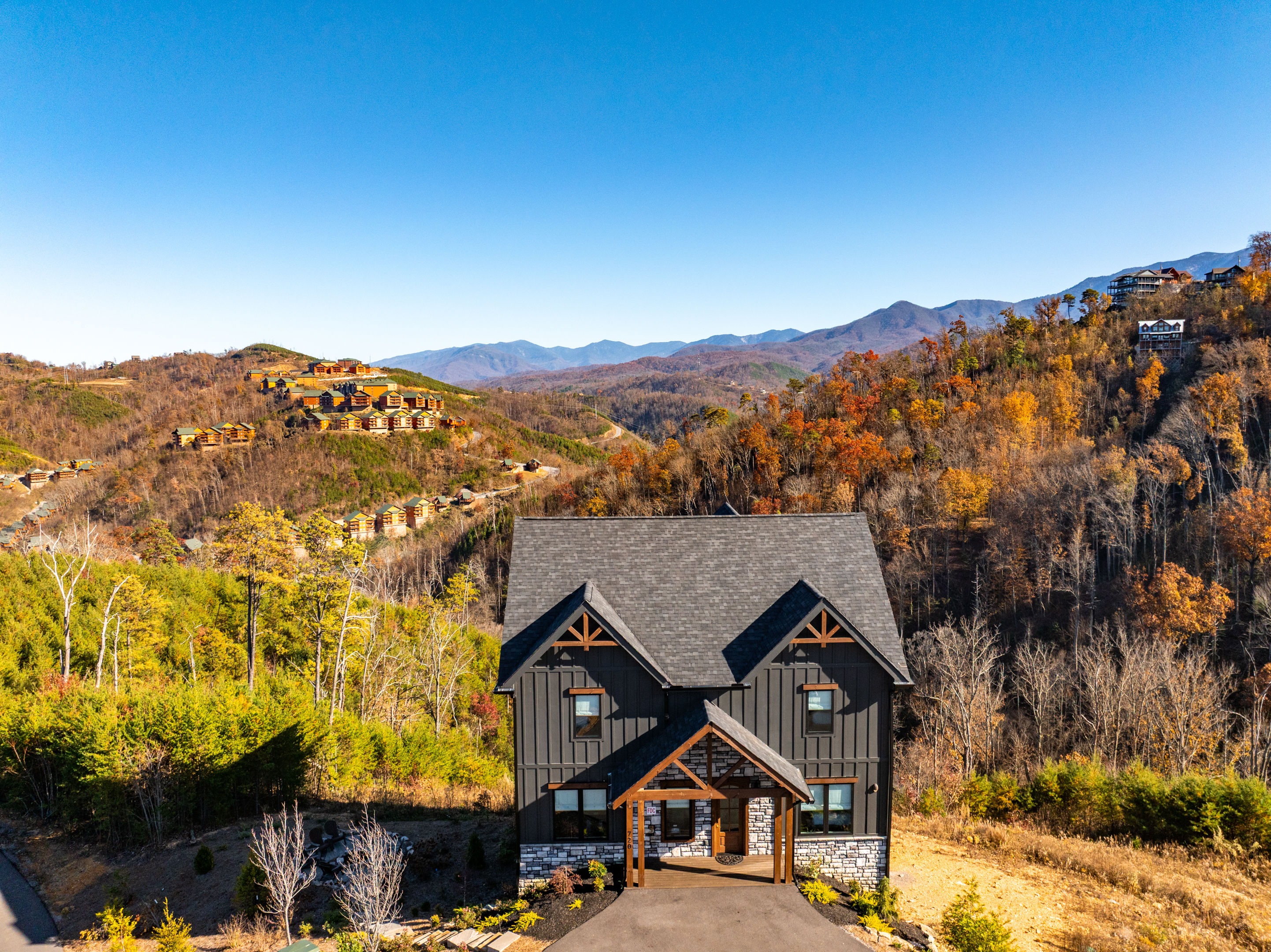 Aerial view of a modern mountain cabin with dark siding and stone base overlooking fall‑colored hills and distant blue mountain ridges under a clear sky, with clusters of hillside vacation homes visible