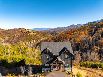 Aerial view of a modern mountain cabin with dark siding and stone base overlooking fall‑colored hills and distant blue mountain ridges under a clear sky, with clusters of hillside vacation homes visible