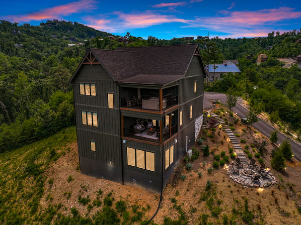 Three-story dark mountain cabin on a wooded hillside at sunset, with covered balconies, warm lit windows, a winding path and stone fire-pit seating area overlooking a forested valley.