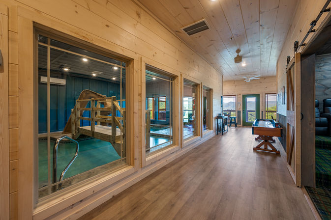 Sunlit wooden hallway in a rustic cabin with windows into an indoor pool featuring a wooden bridge and slide, a game table, and a balcony door with distant mountain views.