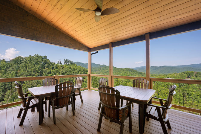 Inviting covered wooden deck with ceiling fan, two outdoor dining tables and chairs overlooking lush forest and rolling mountain vista under a clear blue sky.