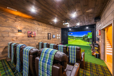 Cozy rustic wood-paneled basement home theater with indoor golf simulator, projector screen showing a golf course, leather recliners draped in blue-green plaid throws and putting-green turf with golf clubs.