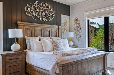 Cozy modern farmhouse bedroom with a rustic wooden bed dressed in white and patterned pillows and a chunky throw, matching wood nightstands with white ceramic lamps, black shiplap accent wall with gold circular wall art, and a large window with leafy outdoor view.