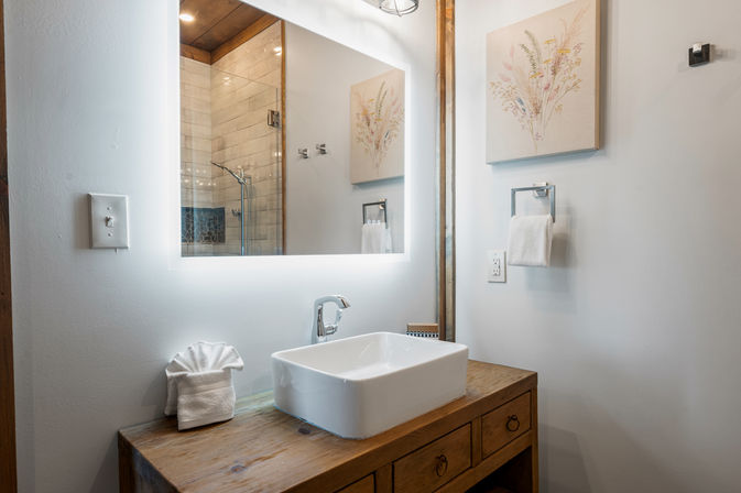 Cozy modern rustic bathroom vanity with wooden countertop, white rectangular vessel sink, chrome faucet, illuminated LED mirror, folded towel and tiled shower reflected in the mirror