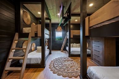 Cozy rustic mountain cabin bunk room with multiple wooden bunk beds and ladders, dark plank walls, round woven rug, rattan pendant light and a window framing a scenic mountain view.