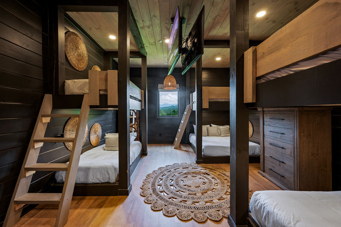 Cozy rustic mountain cabin bunk room with multiple wooden bunk beds and ladders, dark plank walls, round woven rug, rattan pendant light and a window framing a scenic mountain view.
