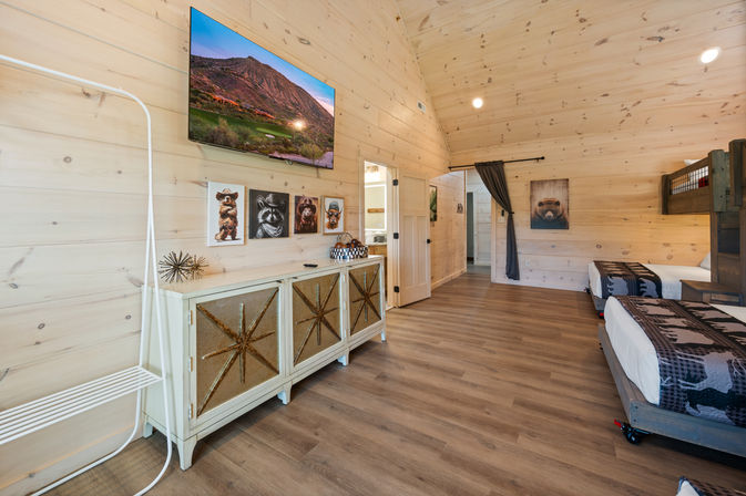 Spacious rustic cabin bedroom with light pine walls and vaulted ceiling, wood-look floors, twin and bunk beds with bear-pattern bedding, wall-mounted TV showing a mountain scene, animal artwork above a star-panel cabinet — bright family-friendly vacation rental interior
