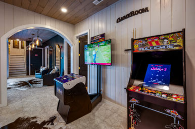 Cozy rustic home game room with wood-paneled walls and ceiling, retro arcade cabinet and cocktail arcade table, wall-mounted TV showing a golf game, cowhide rugs, and arched entry to a seating nook with pendant lights.