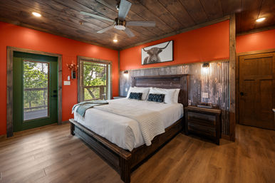 Rustic cabin bedroom with king bed in white linens, corrugated metal accent wall, warm orange walls and wood-plank ceiling with fan, wooden nightstands and a green door opening to a tree-lined balcony, framed highland cow print above the headboard.