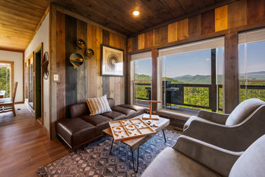 Cozy modern-rustic living room with wood-paneled walls, leather sofa and armchairs, coffee table with tic-tac-toe, and large windows opening to a green mountain view and balcony.