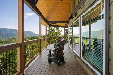 Covered mountain-cabin porch with wooden deck and Adirondack chairs overlooking a lush forest and panoramic mountain valley view, glass windows reflecting the scenery.