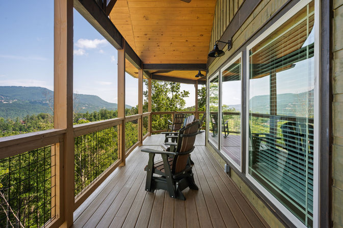 Covered mountain-cabin porch with wooden deck and Adirondack chairs overlooking a lush forest and panoramic mountain valley view, glass windows reflecting the scenery.