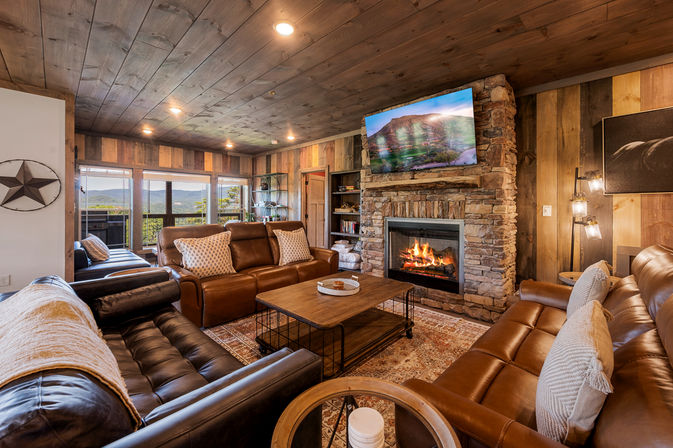 Cozy rustic mountain cabin living room with wood-paneled ceiling and walls, stone fireplace with roaring fire and TV above, leather sofas, coffee table, and large windows framing scenic mountain views.