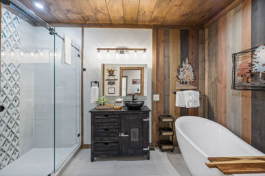 Cozy rustic-modern bathroom with wood-paneled walls and ceiling, glass walk-in shower with geometric tile accent, dark wood vanity with black vessel sink, and white freestanding tub with wooden caddy.
