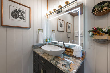 Bright, cozy farmhouse-style bathroom vanity with LED-lit mirror and brass sconces, white vessel sink on a multicolored granite countertop, dark gray cabinets, shiplap walls, framed art and decorative floral shelves.