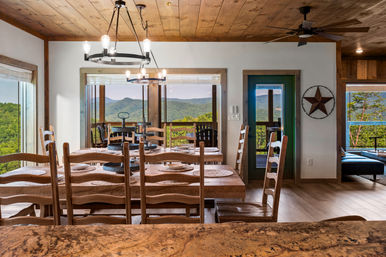 Rustic wood‑paneled mountain cabin dining room with long wooden table and ladder‑back chairs, modern pendant lights, and panoramic windows and door opening to a deck with rolling green hills and distant blue mountains.