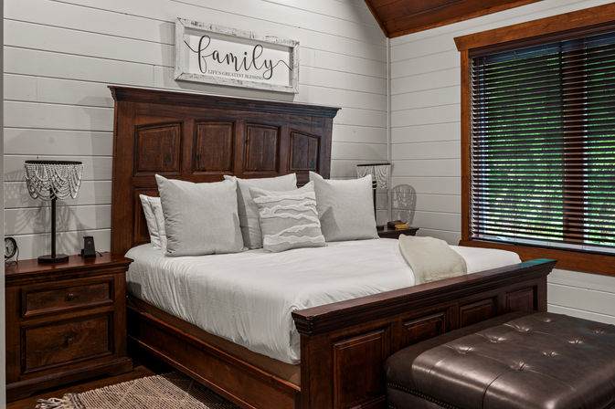 Cozy farmhouse bedroom with dark wooden bed and headboard, white shiplap walls, neutral pillows and linens, beaded bedside lamps, leather ottoman and wooden blinds revealing green foliage outside.