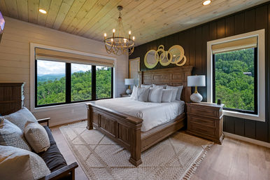 Cozy rustic bedroom with a large wooden king bed dressed in white linens, chandelier overhead, circular mirrors above the headboard, matching nightstands and lamps, and two large windows framing lush green forest and distant mountain views.