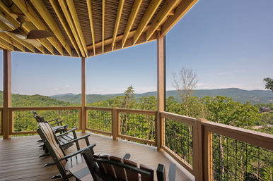 Covered wooden deck with Adirondack chairs overlooking rolling green mountains and forest, panoramic mountain view under a clear blue sky.