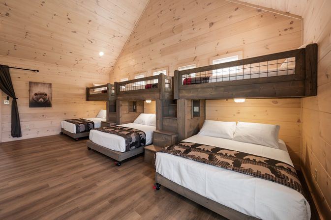 Cozy mountain cabin bunk room with vaulted pine walls and wood floors, three rustic bunk-bed units with lower beds and upper loft bunks, bear-print throws, soft lighting and simple decor ideal for family or group stays.