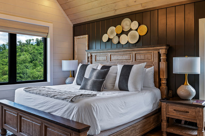 Cozy rustic-modern bedroom with a king-size wooden bed, white linens and gray accent pillows, matching nightstands and lamps, circular wall art above the headboard, and a large window framing lush green forest and mountain views.