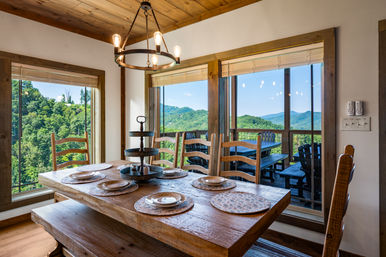 Cozy rustic wooden dining room with a farmhouse table set for six, large windows revealing lush mountain views and a deck with outdoor seating.
