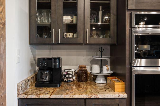 Modern home kitchen coffee station on speckled granite countertop with a black Keurig machine, wire basket labeled 'COFFEE', brown ceramic jar, two-tier white serving stand, dark glass-front cabinets and stainless built-in oven