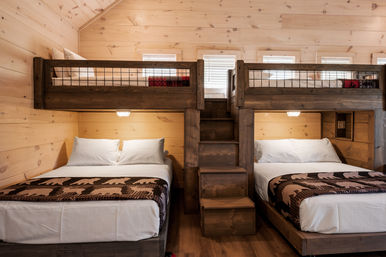 Cozy rustic mountain cabin bedroom with wooden bunk bed unit, two double beds below and loft bunks above connected by central stairs, white linens and patterned bear blankets, pine plank walls and small windows.