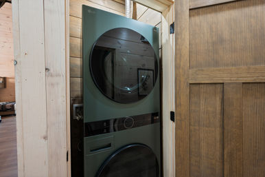 Stacked green LG washer and dryer with sleek round glass doors inside a narrow wood-paneled laundry closet behind a barn-style sliding door in a rustic cabin