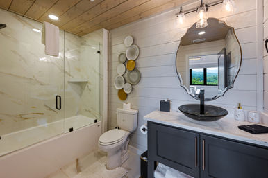 Cozy modern farmhouse bathroom with wood-plank ceiling, glass-enclosed marble tub/shower, white toilet, decorative wall plates, gray vanity with black vessel sink and scalloped mirror reflecting a mountain view.