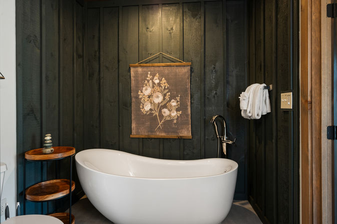 Freestanding white bathtub in a cozy rustic bathroom with dark vertical wood paneling, floral wall hanging, chrome freestanding faucet, wooden tiered side table with stacked stones and folded white towels.