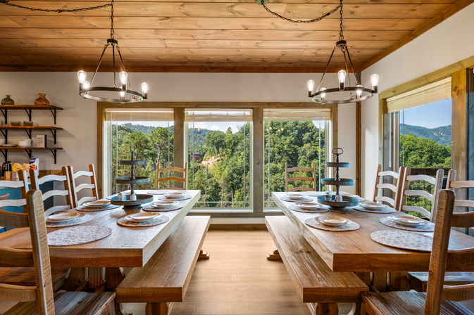 Cozy rustic dining room with wood-paneled ceiling, twin long wooden tables and benches set with plates beneath industrial chandeliers, facing large panoramic windows with a lush forested mountain view.