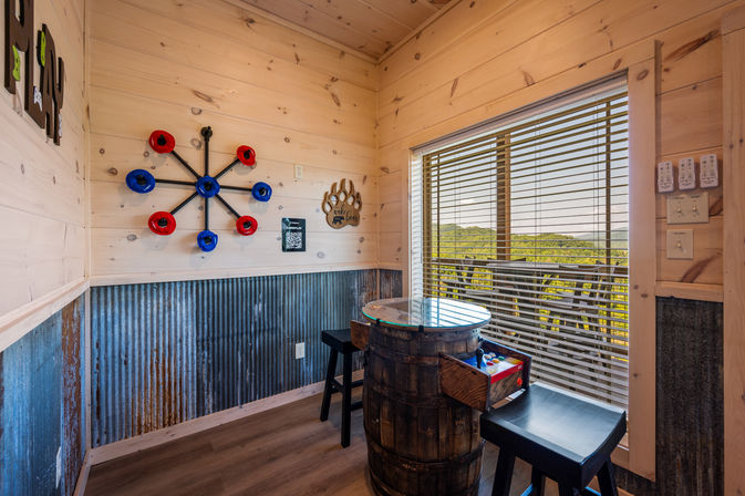 Rustic mountain cabin game nook with light wood walls, corrugated metal wainscoting, barrel arcade table and stools, wall-mounted ring-toss game, and a large window with blinds framing green hill views.