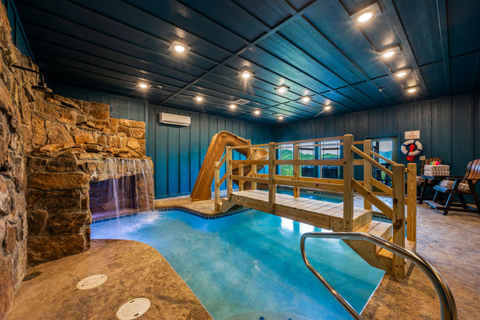 Indoor pool with turquoise water, rustic rock waterfall, tan waterslide and wooden footbridge in a blue-paneled recreation room with lounge seating and life ring.