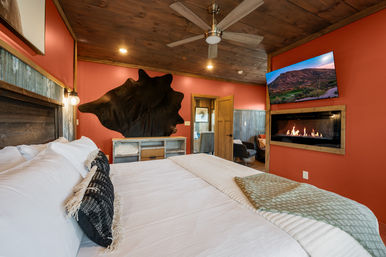 Cozy rustic lodge bedroom with king bed and white linens, wooden ceiling with fan, red accent walls, wall-mounted TV above a linear gas fireplace, and large decorative hide on the wall.