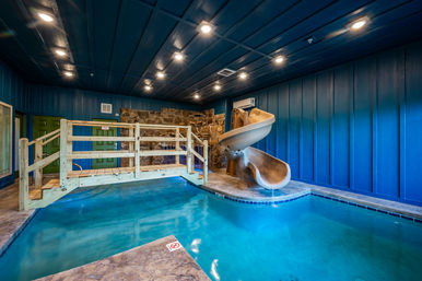 Indoor resort-style pool with turquoise water, tan spiral water slide, small wooden bridge, blue paneled walls, stone accent wall and recessed ceiling lights