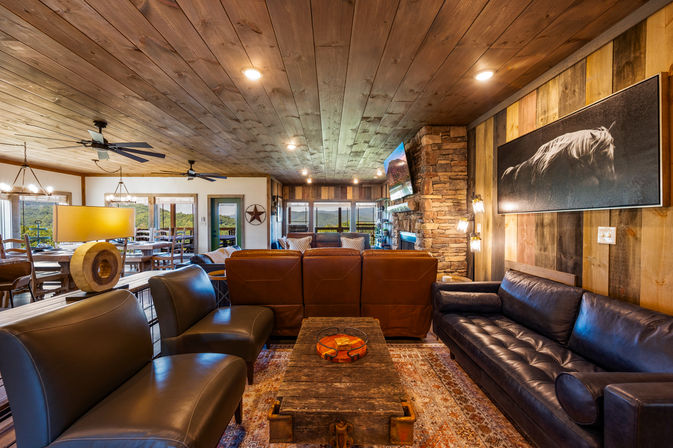 Cozy rustic lodge living room with leather sofas and chairs, reclaimed wood ceiling and plank walls, stone fireplace with mounted TV, wooden coffee table on a patterned rug, and dining area with panoramic windows overlooking forested hills.