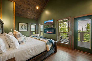 Cozy rustic mountain cabin bedroom with green walls, wood-beamed ceiling, queen bed, wall-mounted fireplace and balcony with forest views.