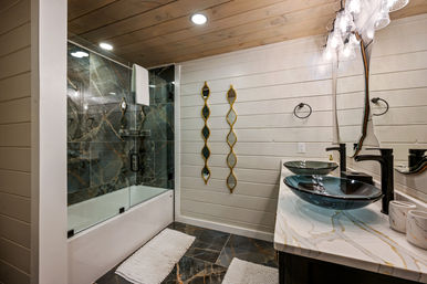 Modern rustic bathroom with glass-enclosed tub-shower and dark marble tiles, white wood plank walls, double glass vessel sinks on a marble countertop and gold decorative mirrors