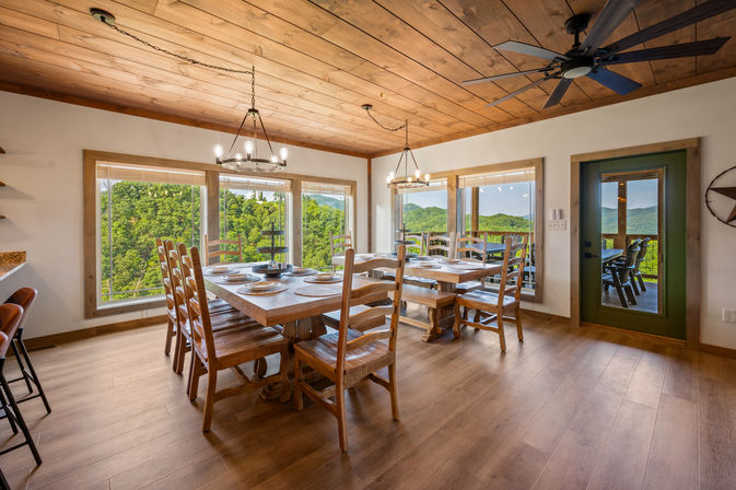 Sunlit rustic mountain-cabin dining room with a long wooden farmhouse table set for eight, chandeliers and ceiling fan, large windows framing green forested hills and a deck with outdoor seating.