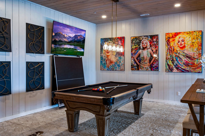 Stylish indoor game room with wooden pool table and black felt beneath a modern multi-bulb pendant, wall-mounted flat-screen TV, decorative metal wall panels, and three colorful pop-art portraits of blonde women on white shiplap walls