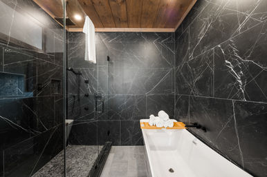 Spa-like modern bathroom with black marble walls, glass-enclosed shower, freestanding white bathtub with wooden caddy and rolled towels, and warm wood ceiling