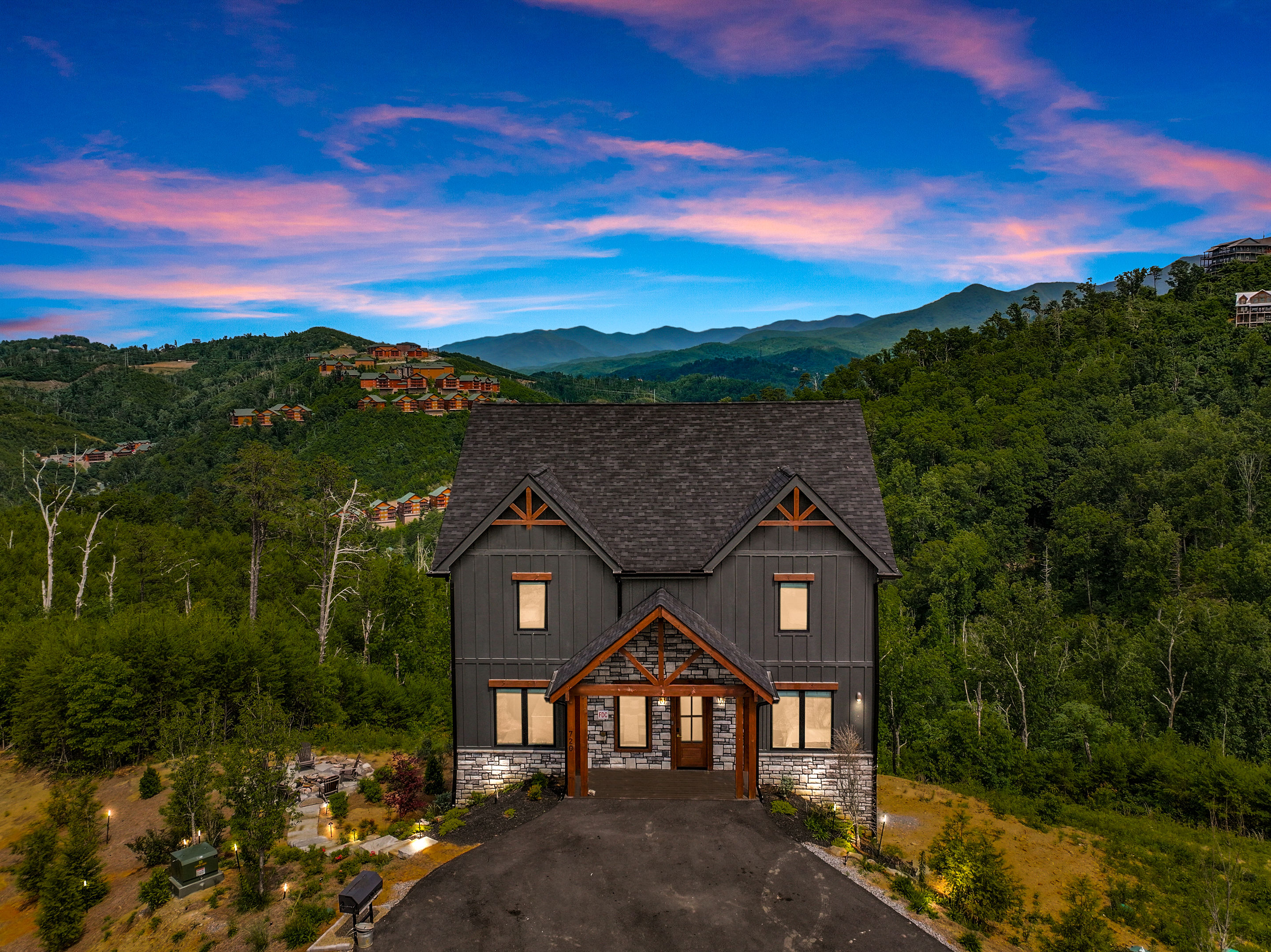 Modern two-story mountain cabin with wood and stone accents perched above a forested valley, colorful pink-and-blue sunset sky and distant mountain ridges