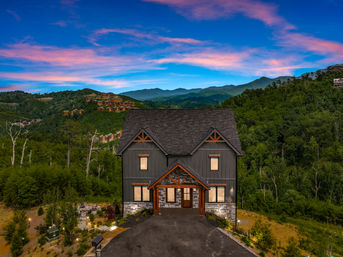 Modern two-story mountain cabin with wood and stone accents perched above a forested valley, colorful pink-and-blue sunset sky and distant mountain ridges