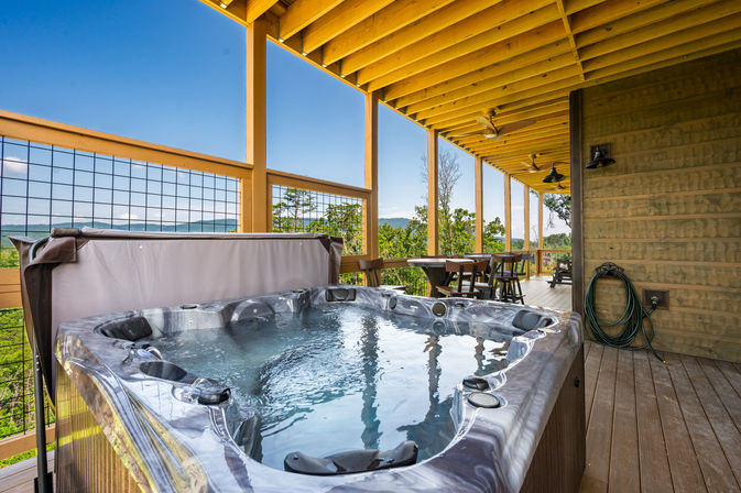 Covered wooden deck with a bubbling hot tub and outdoor dining set overlooking forested hills and distant mountain views under a bright blue sky