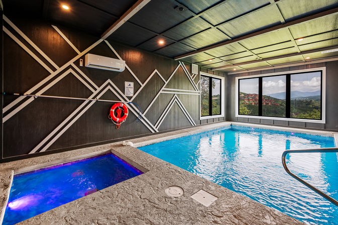 Inviting indoor swimming pool and glowing blue hot tub in a contemporary lounge with geometric accent wall, life preserver, and large windows framing rolling mountain views.