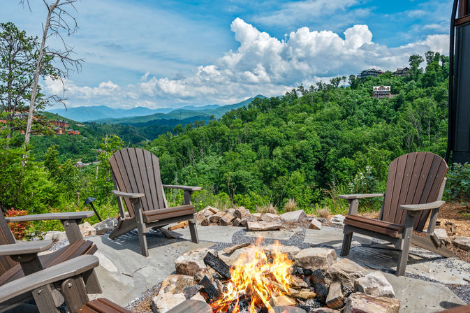 Cozy outdoor fire pit surrounded by Adirondack chairs on a mountain overlook, lush green forested valley and blue ridgelines under dramatic summer clouds